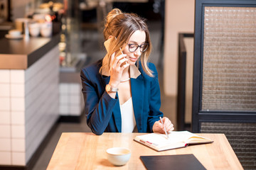 Young businesswoman strictly dressed in suit talking with phone working at the modern cafe interior