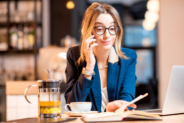 Disappointed businesswoman strictly dressed in the suit talking phone while working with laptop at the modern cafe interior