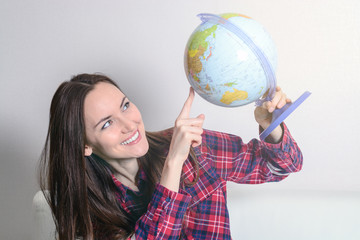 Fototapeta premium Happy young brunette woman dreams about traveling on vacation with a globe in hands on a white sofa in the room