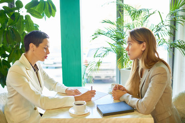Side view portrait of two modern young women discussing work sitting at table in cafe during business meeting or job interview