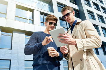 Low angle portrait of two successful young people using digital tablet outdoors standing against modern office building, copy space