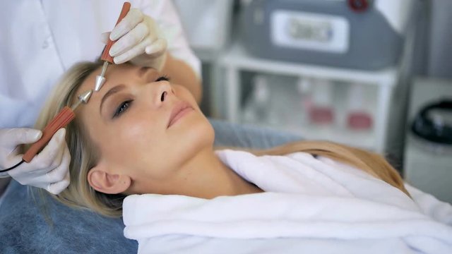 Close-up of beautiful woman receiving facial microcurrent treatment from therapist at beaty salon