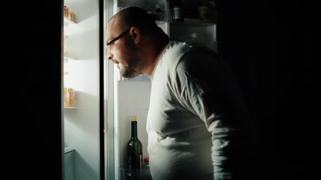 Fat man opens a refrigerator at night and is looking for something to eat, night hunger, gluttony concept