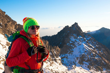 Smiling attractive young woman hiking in winter high mountains with backpack, she looking at the peak of mountain at sunset.