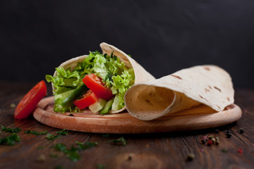  burrito with greens, cucumbers, tomatoes and sausages on a round wooden board on a wooden background