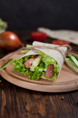  burrito with greens, cucumbers, tomatoes and sausages on a round wooden board on a wooden background
