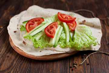  burrito with greens, cucumbers, tomatoes and sausages on a round wooden board on a wooden background