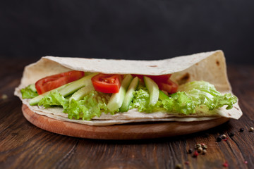  burrito with greens, cucumbers, tomatoes and sausages on a round wooden board on a wooden background