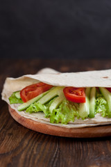  burrito with greens, cucumbers, tomatoes and sausages on a round wooden board on a wooden background