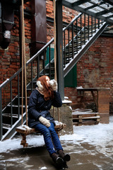 red-haired girl is drinking coffee on  wooden swing in winter street