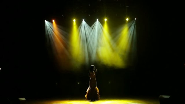A beautiful girl vocalist performs a song on the stage lit with multi-colored searchlights. Beautiful girl sings a song on illuminated stage.