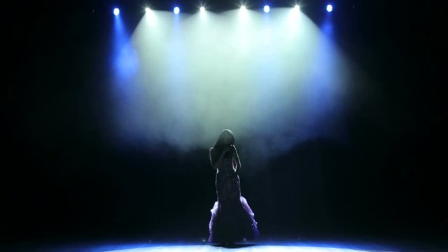 A young woman in a luxurious evening dress sings a song and dances on a dark smoky stage.