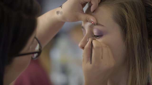 Makeup artist is applying eyeliner on eyelid of lady in beauty salon. Young expert puts neatly black liquid color on eyes of beautiful model, pressing on skin with finger. Talented woman in glasses