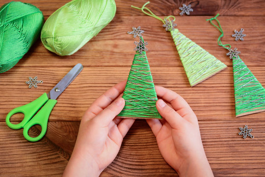 Kid is holding a Christmas tree decoration in his hands. Kid is showing a Christmas tree decoration. Merry Christmas tree project for kids. Scissors, green cotton yarn on a wooden table 