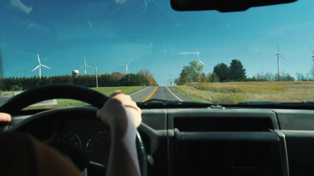 The driver at the wheel of the car goes on a rural road. On the sides there are many wind turbines. Ecotourism and clean energy