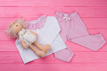 Doll and pajama set on pink wooden table. Top with cute collar and pants with drawstring. Little girl's toys and clothes.