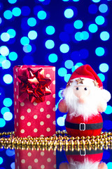Santa Claus toy, gift in package and golden beads on a glass table with a beautiful blue bokeh - 184251531