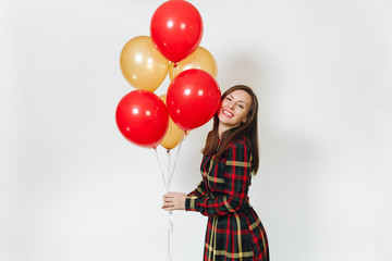 Beautiful caucasian young happy woman in long plaid checkered dress with shy charming smile, red, yellow golden balloons, celebrating birthday, on white background isolated. Holiday, party concept.