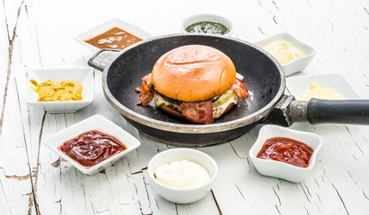 Hamburger in a frying pan with sauces set around on white wooden background with cracked paint.