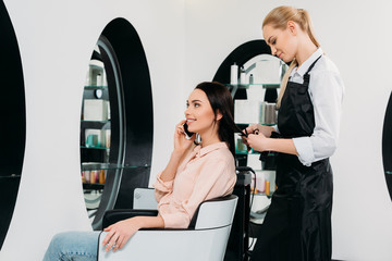 woman talking by smartphone while hairdresser cutting hair