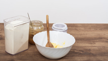 Ingredients for making sweet dough on the kitchen table. Flour, honey, sugar and butter in white bowl on the beautiful wooden background.