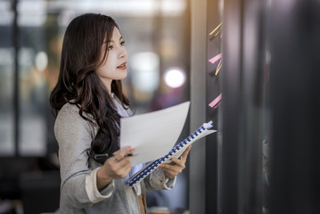 Young asian business woman holding paper and looking at sticking colourful notes on glass wall