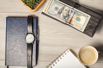 Black leather wallet with dollar bills, notebook, men's watch, pen, cup of coffee and houseplant in a pot