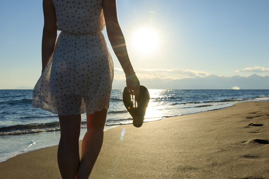 Young, sexy woman is walking at a beautiful sunset beach. holding her flip flops in her hand. sexy short dress skirt. wet feet legs. sundown mountains