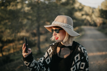 Cute woman in sunglasses taking a photo with a mobile phone in nature at sunset.
