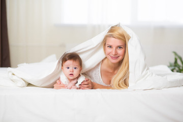 Obraz na płótnie Canvas Portrait of young happy smiling mother with infant baby girl liying on the bed covered with a white blanket. Happy family concept