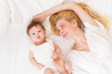 Obraz na płótnie Canvas Happy loving family concept. Beautiful mother playing with her baby girl in the bedroom. They smiling and hugging together on white bed linens, top view