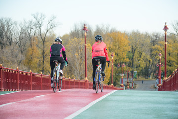 Two Young Female Cyclists Riding Road Bicycles on Bridge Bike Line in Cold Autumn Day. Healthy Lifestyle Concept.