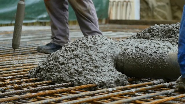 Slow motion shot: Liquid concrete pouring from tube on armature reinforcement bar, and worker using concrete vibrator machine behind.
