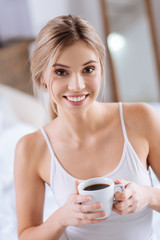 Enjoy this drink. The portrait of a charming slender young woman holding a cup of coffee and smiling at the camera