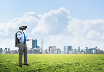 Fototapeta na wymiar Camera headed man standing on green grass against modern cityscape