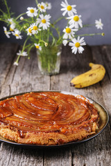 Obraz na płótnie Canvas Traditional homemade banana caramel pie on vintage wooden table decorated a bouquet of chamomiles. Upside down banana cake. Selective focus 