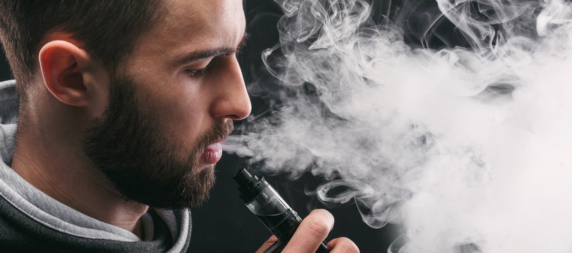 Young man vaping e-cigarette with smoke on black