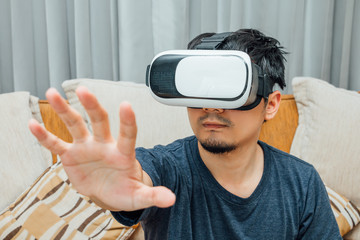 Asian man wearing virtual reality glasses expressing on brown sofa, smartphone using with VR glasses