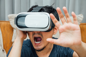Asian man wearing virtual reality glasses expressing emotion panic on brown sofa, smartphone using with VR glasses