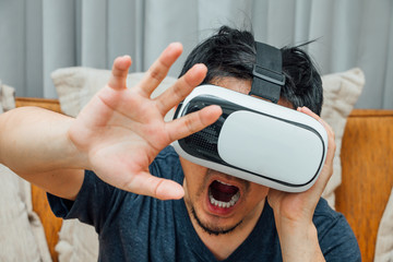 Asian man wearing virtual reality glasses expressing emotion panic on brown sofa, smartphone using with VR glasses