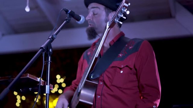 Closeup of guitarist playing resonator and singing at an outdoor concert at a restaurant at night.