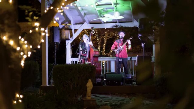 Moving through the trees with lights looking at the blues band performing at an outdoor cafe at night.