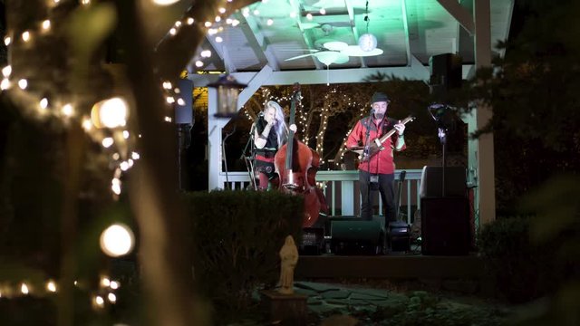 Moving through the trees with lights looking at the blues band performing at an outdoor cafe at night.