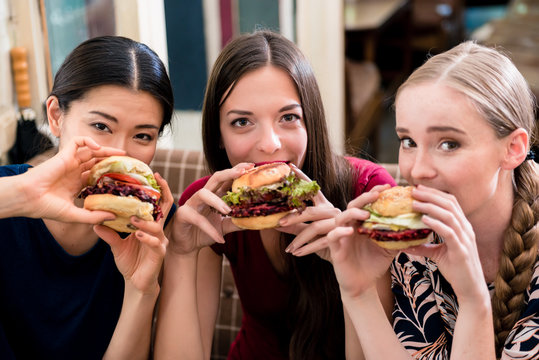 Portrait of three young women looking at camera while eating hamburgers indoors