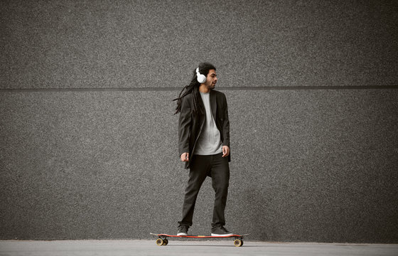 Close up of stylish handsome happy dreadlocks skater man in a suit standing on skate while listening music on the street.