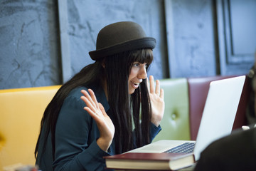 Giovane studentessa con cappello a bombetta guarda il computer incredula con le mani alte seduta in un divano colorato.