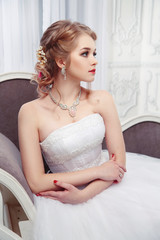 Fototapeta na wymiar A sweet bride in a beautiful dress sits in an armchair in a chic, light interior.