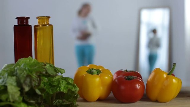 Blurry woman admiring her shape in the mirror in domestic room with fresh coolorful vegetables on foreground. Cheerful girl looking at reflection of her slimmed figure in the mirror. Healthy lifestyle