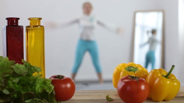 Blurred toned young fit woman doing warm up exercises in domestic interior with healthy food on foreground. Sporty girl in sportswear working out at home. Healthy eating, dieting and fitness concept.