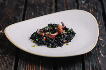 Black risotto with cuttlefish ink and octopus tentacles on irregular shape plate on dark wood background